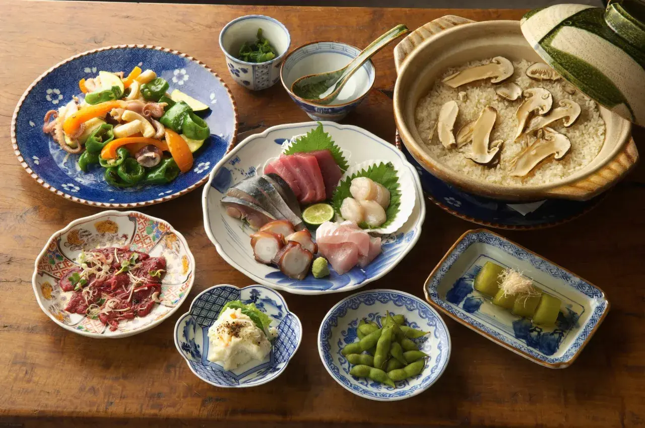A spread of Japanese side dishes, including sashimi, edamame, potato salad, and a vegetable stir-fry, complements a pot of rice with mushrooms.