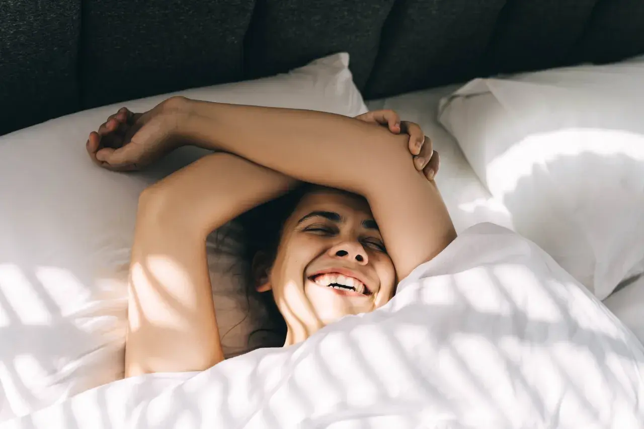 Glückliches Lachen im Bett, Arme über dem Kopf verschränkt. Ein Gefühl von Freiheit, vielleicht ohne Kissen schlafen.