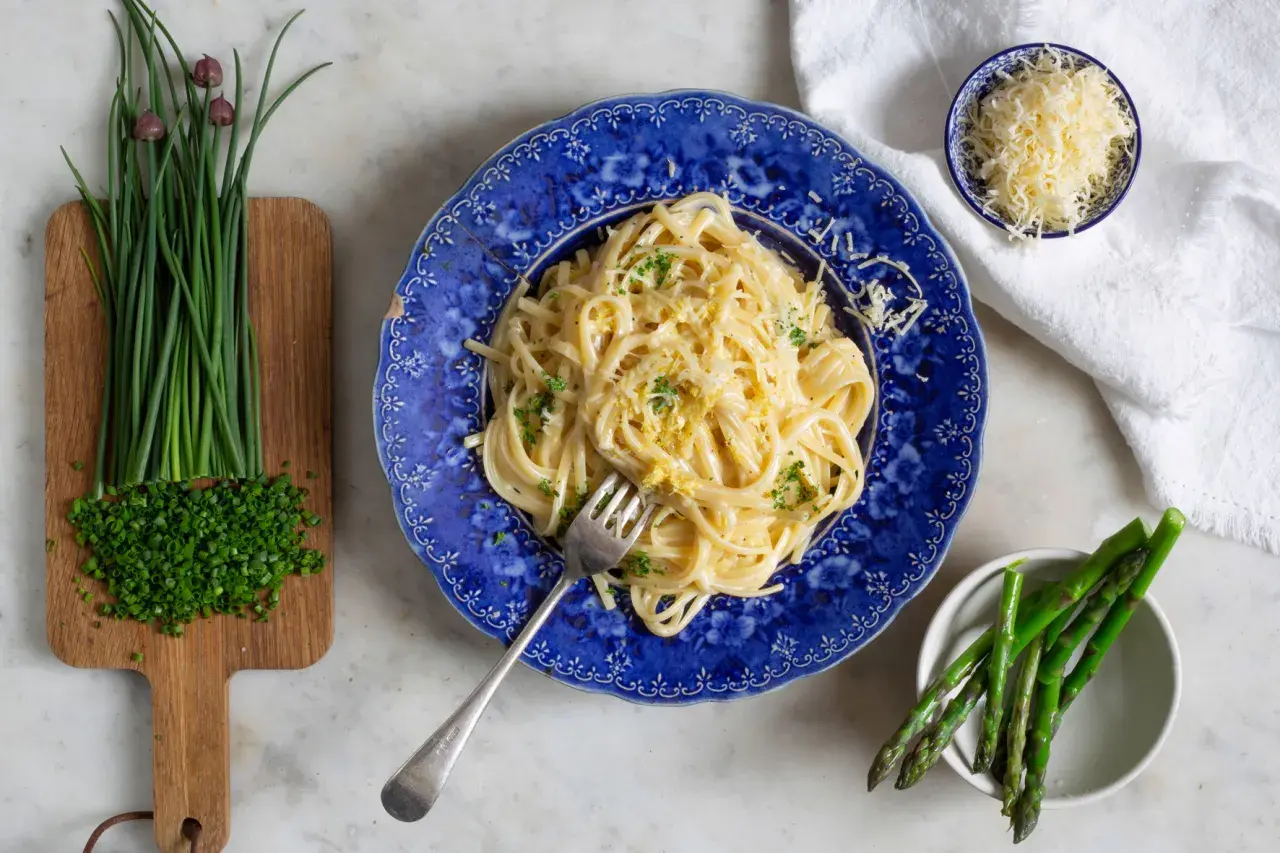 En tallrik krämig pasta limone, toppad med riven ost och persilja. Bredvid finns färsk gräslök och sparris.