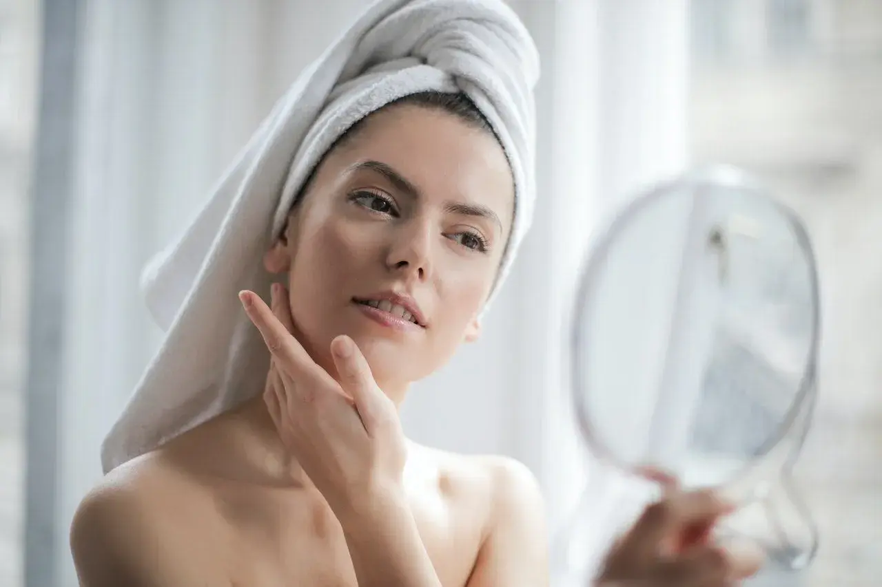 Donna con asciugamano in testa si guarda allo specchio, la sua pelle luminosa grazie all'acido ialuronico e vitamina C insieme.