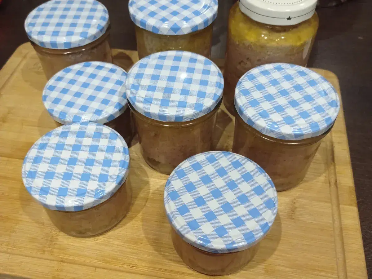 Mehrere Gläser mit selbstgemachter Leberwurst, verschlossen mit blau-weißen Karodeckeln, stehen auf einem Holzbrett.