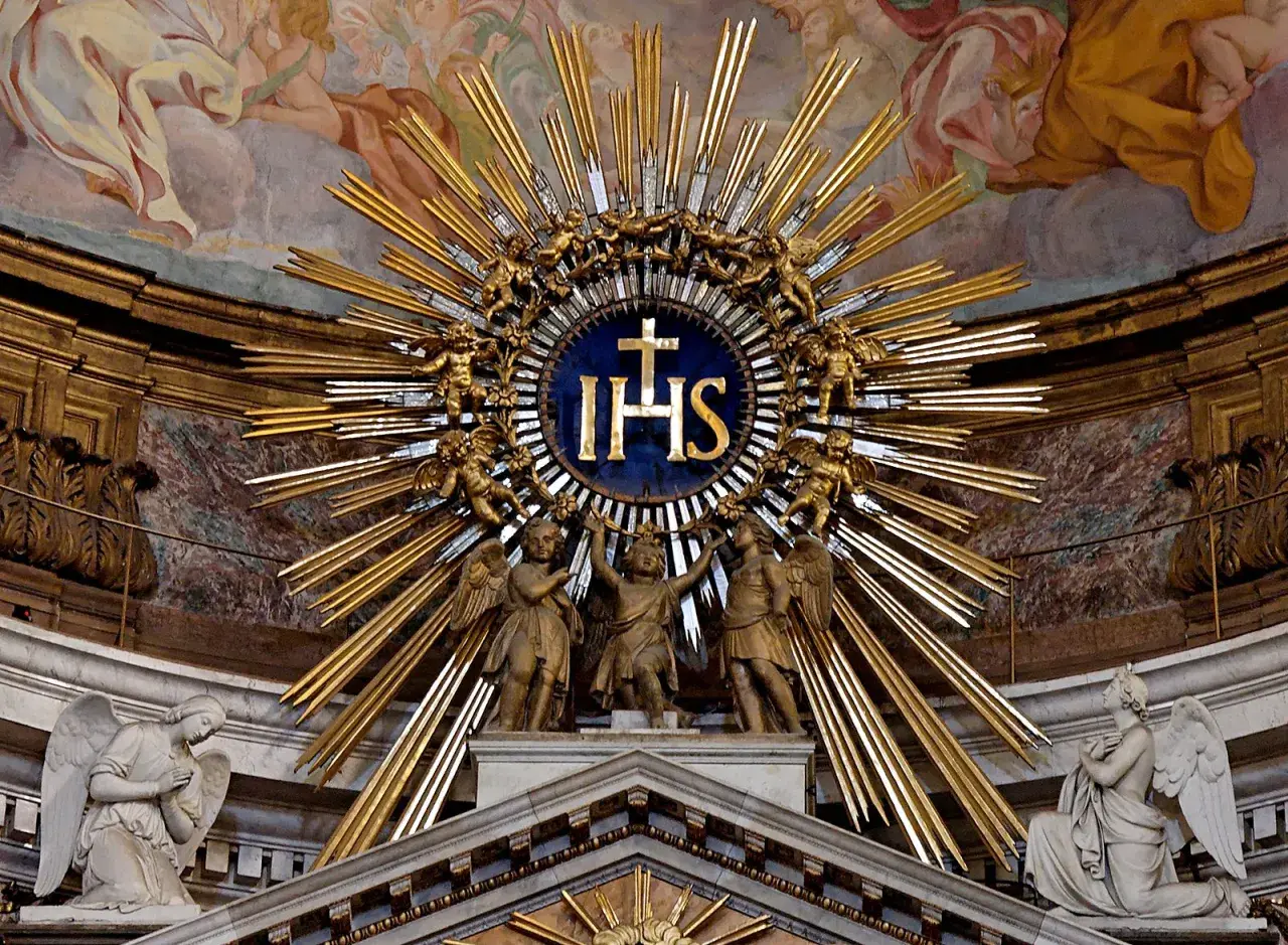 Goldene Strahlenkrone mit IHS-Symbol, das für Jesus steht. Putten und Engel umgeben das Symbol, das die göttliche Bedeutung von IHS unterstreicht.