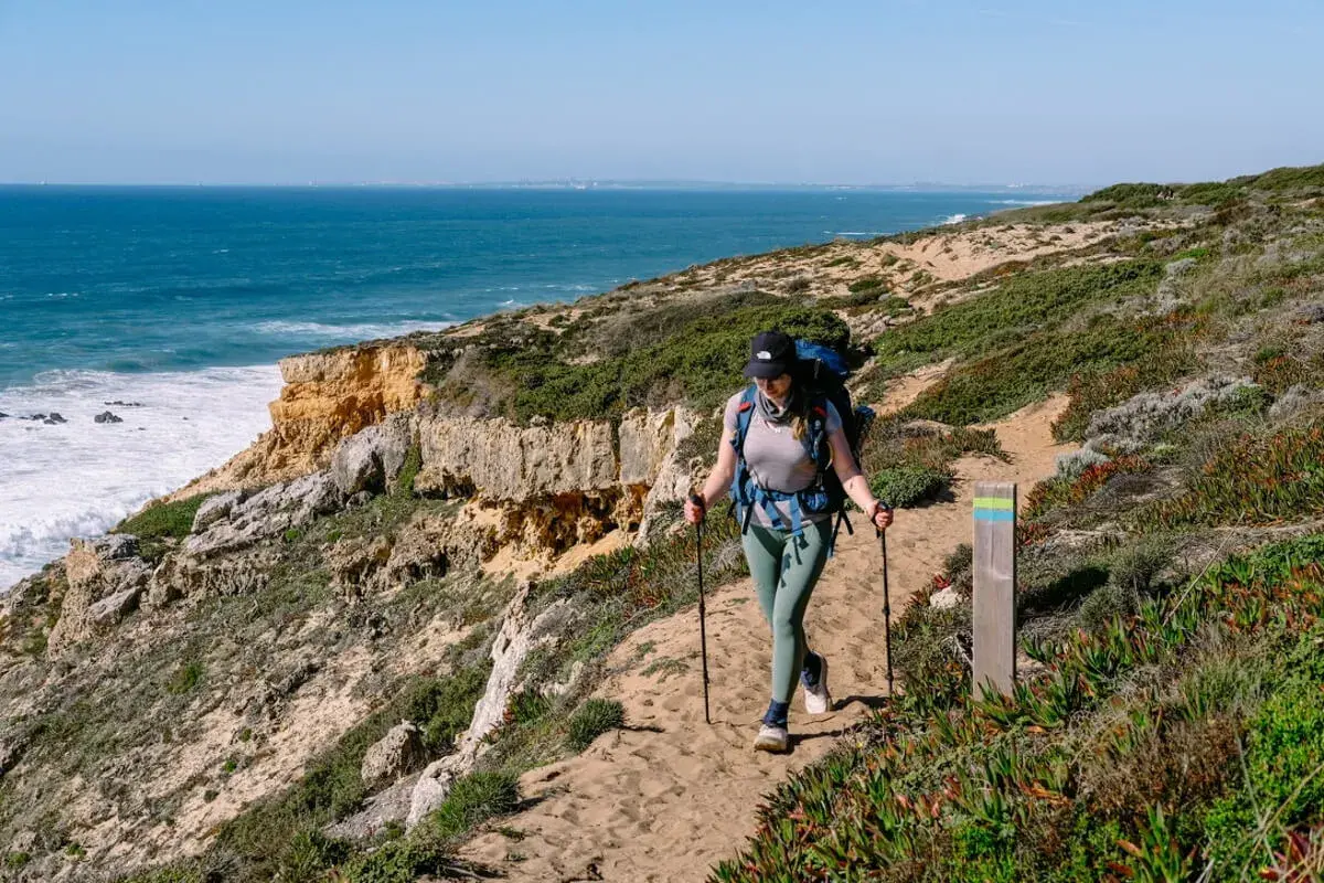 Wanderin auf dem Fisherman's Trail in Portugal. Eine Frau mit Rucksack und Stöcken wandert auf einem sandigen Küstenpfad mit Blick auf das Meer.