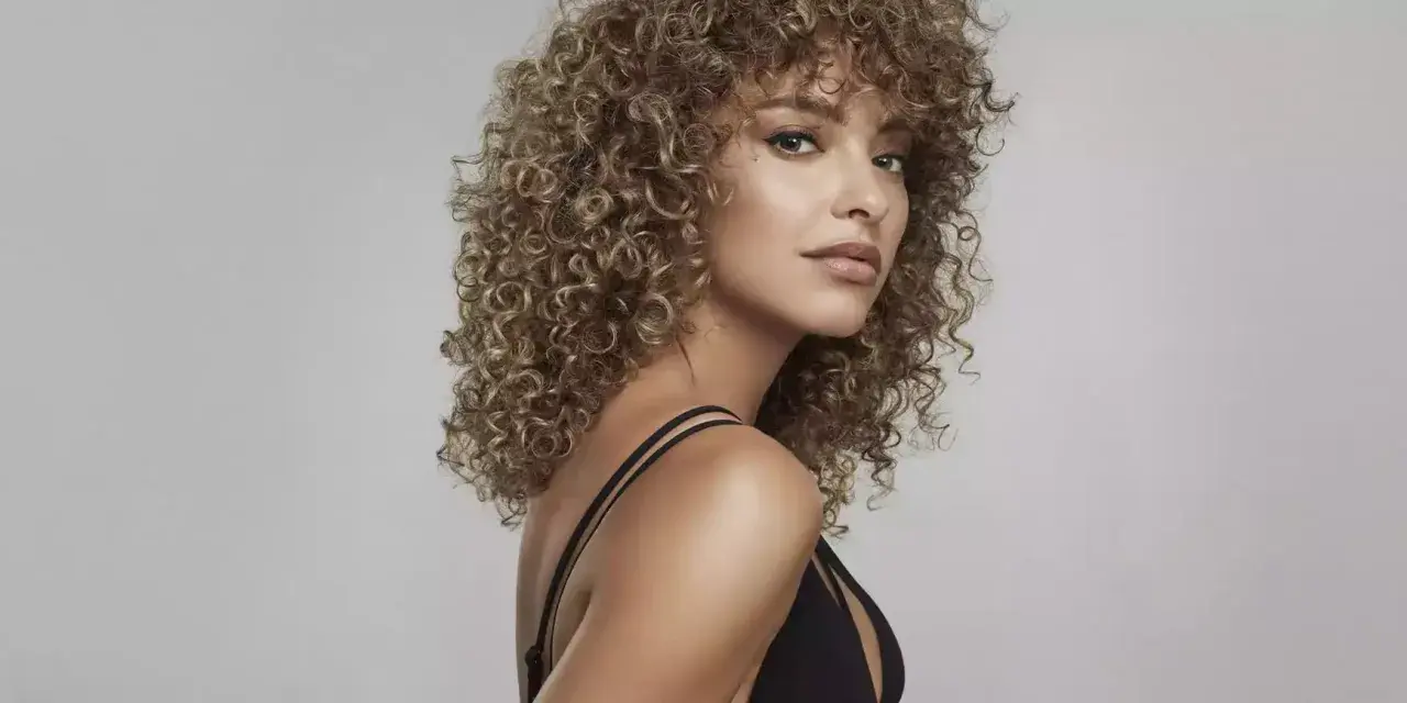 Kobieta z kręconymi, blond grzywkami i w czarnej sukience na jasnym tle.