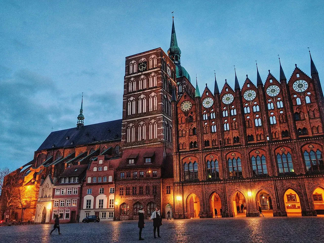 Abendstimmung im **Stralsund Zentrum**: imposante Backsteingotik mit Kirchturm und Rathaus, beleuchteter Marktplatz mit wenigen Passanten.