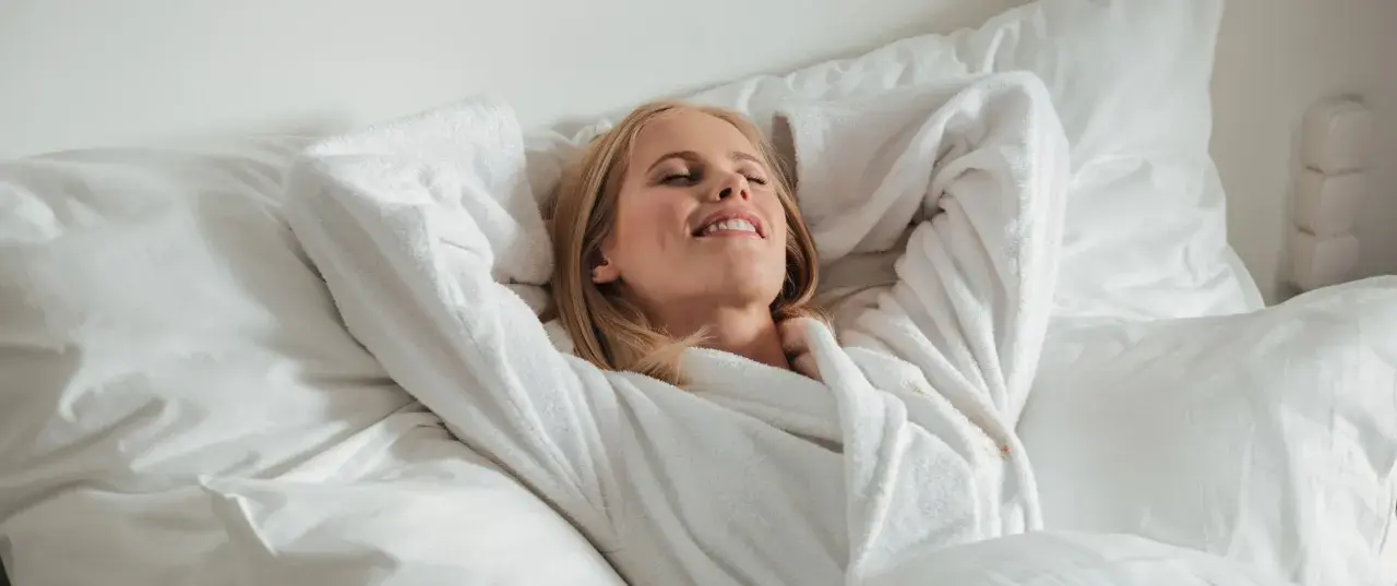 Frau entspannt im Bademantel im Bett, genießt den Moment. Vielleicht denkt sie über die richtige 5-HTP Dosierung nach, um sich noch besser zu fühlen.
