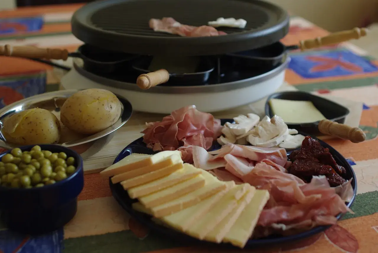 Préparation d'une raclette conviviale : fromage, charcuterie, pommes de terre et petits pois attendent sur la table.
