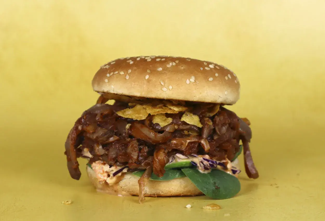 Hamburguesa con pulled pork vegano, cebolla caramelizada y crujientes patatas fritas sobre fondo amarillo.