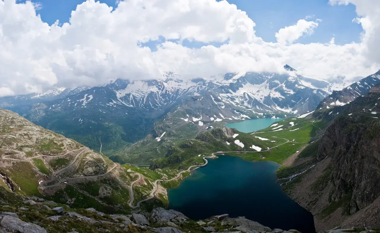 Strada tortuosa sul passo Nivolet, con laghi alpini e montagne innevate sotto un cielo nuvoloso.