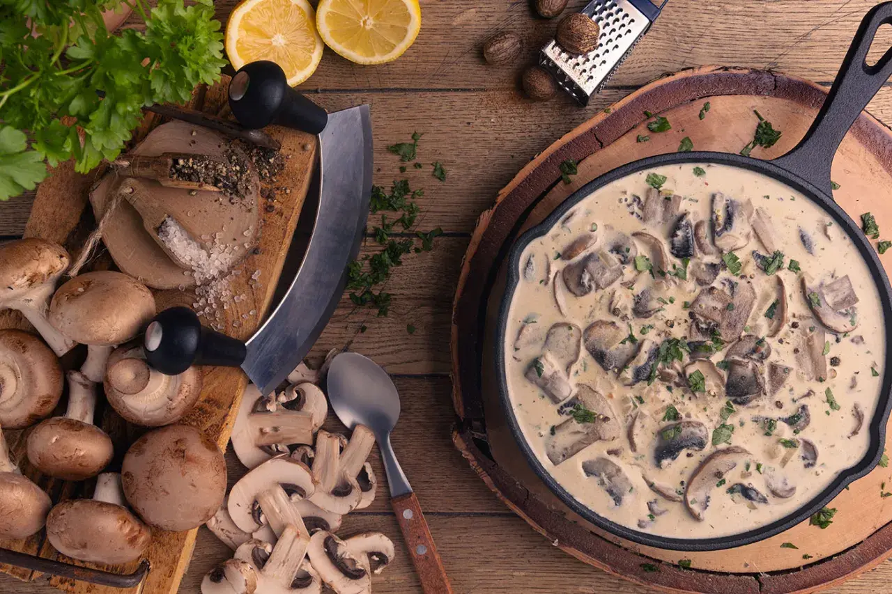 Cremige Rahmchampignons in einer Pfanne, daneben frische Champignons, Kräuter, Zitrone und Gewürze. Ein leckeres Rahmchampignons Rezept zum Nachkochen.
