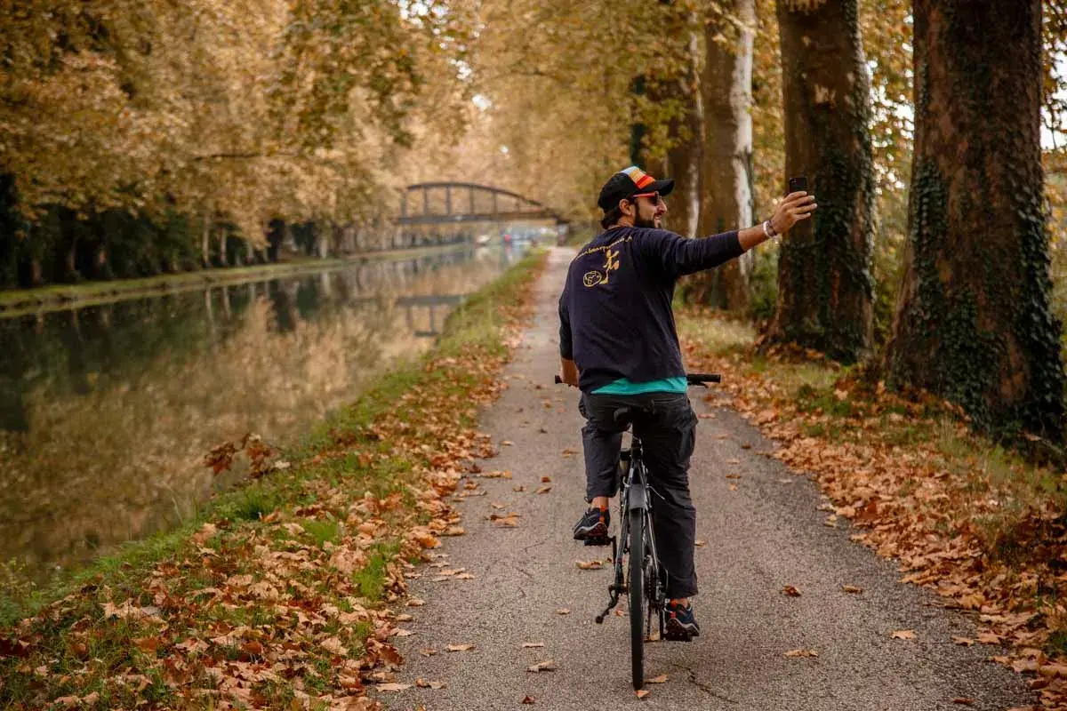 Hombre en bicicleta toma selfie en el Canal du Midi, rodeado de árboles otoñales y hojas caídas.