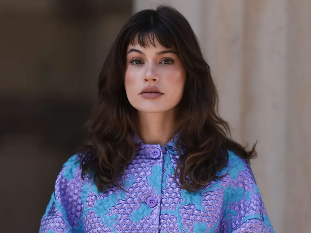 Mujer con melenas cortas con volumen, flequillo y un llamativo abrigo morado y turquesa.