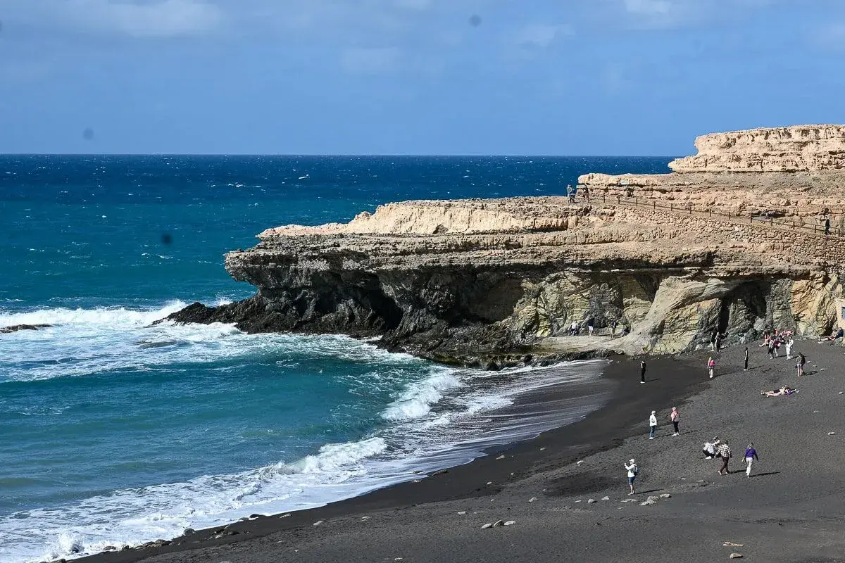Fuerteventura Urlaub Tipps: Schwarzer Strand mit Felsen und türkisblauem Meer. Menschen spazieren und entspannen.