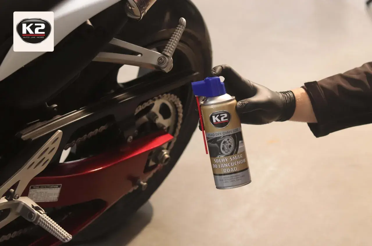 Ręka w czarnej rękawiczce trzyma puszkę smaru do łańcucha K2. Motocykl w tle.