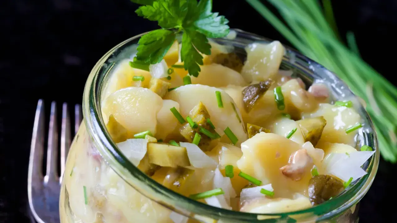Ein köstlicher Kartoffelsalat ohne Mayo, mit Gurkenstücken und frischem Schnittlauch garniert, serviert in einem Glas.
