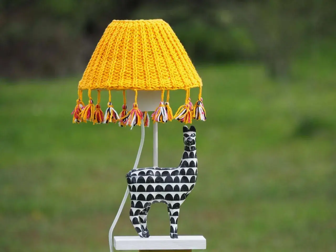 Lámpara techo infantil con pantalla tejida amarilla y borlas de colores. La base es una figura de llama blanca y negra.