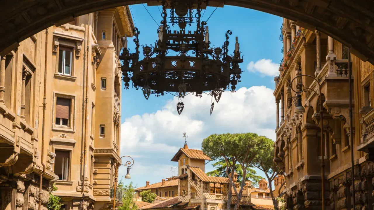 Un imponente lampadario domina la vista del quartiere Coppedè, incorniciato da palazzi storici e un cielo azzurro con nuvole bianche.