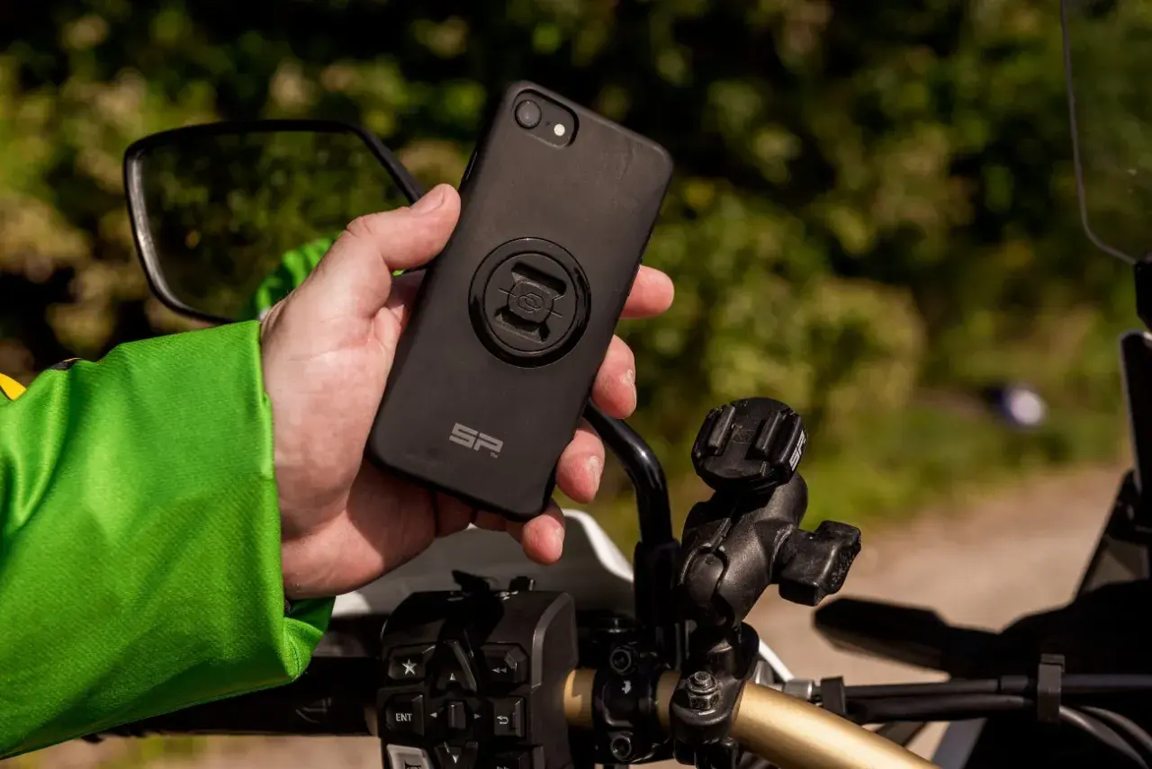 Dłoń trzyma czarne etui na telefon z mocowaniem SP Connect, obok uchwytu na kierownicy motocykla.
