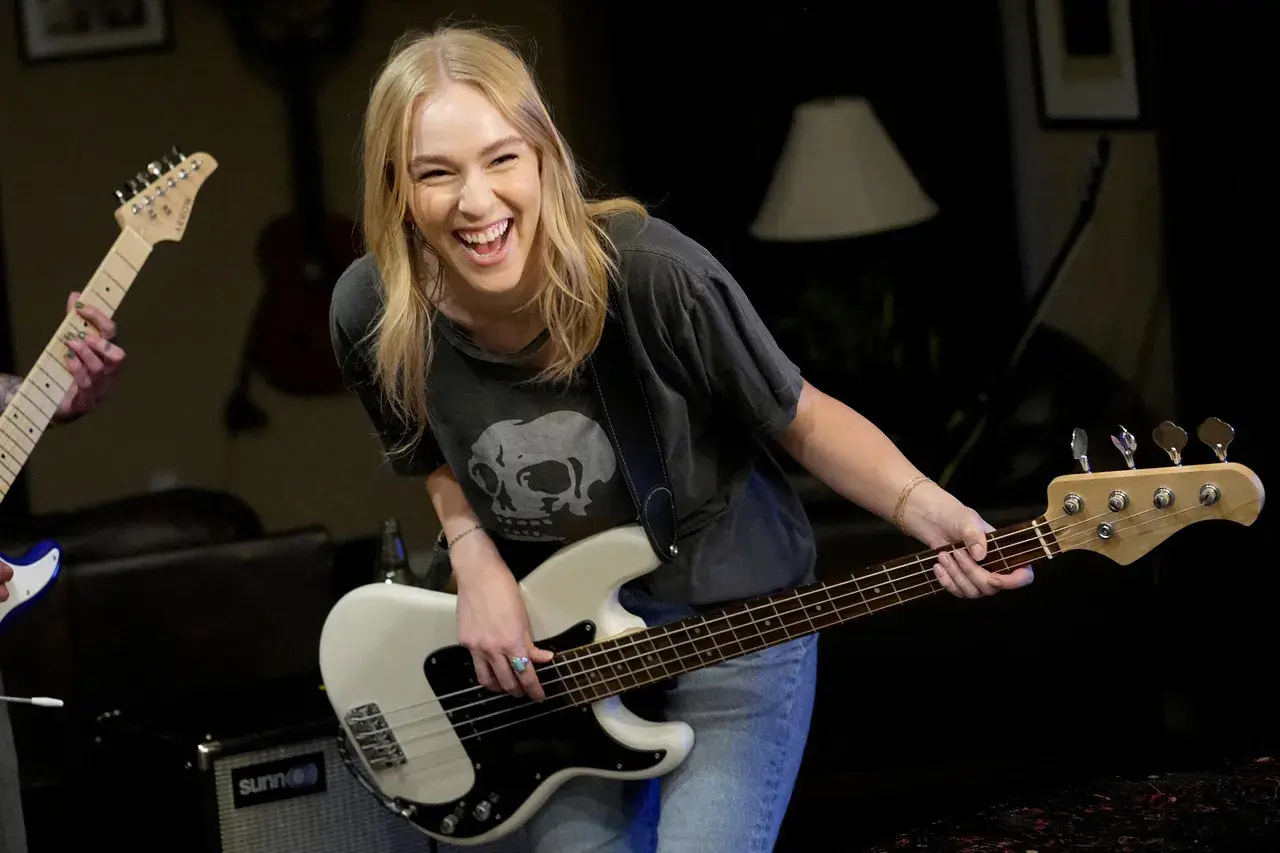 Uśmiechnięta blondynka gra na białym bas instrumencie, w tle inny gitarzysta.