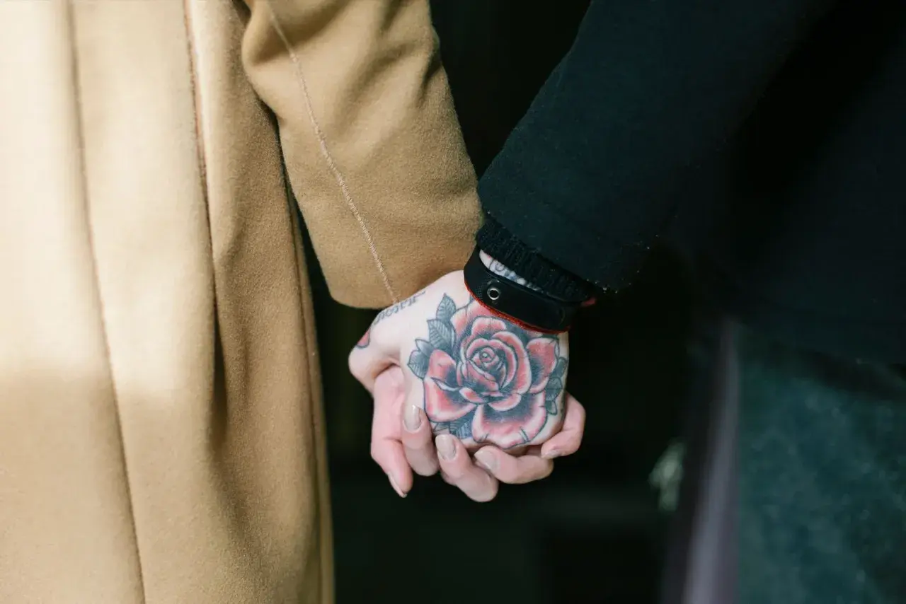 Manos entrelazadas, una con un tatuaje de rosa en fase lechosa, la otra envuelta en un abrigo camel.