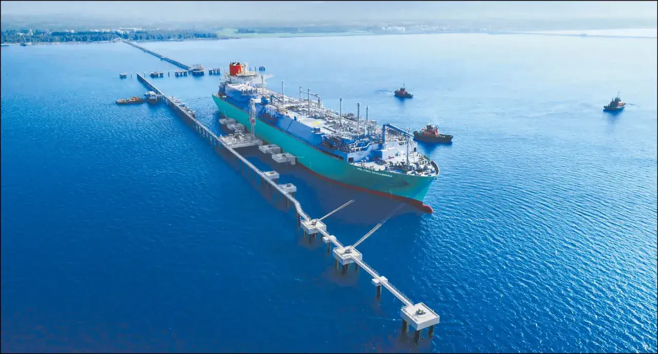 Pływający terminal LNG przycumowany do nabrzeża, z holownikami na wodzie.