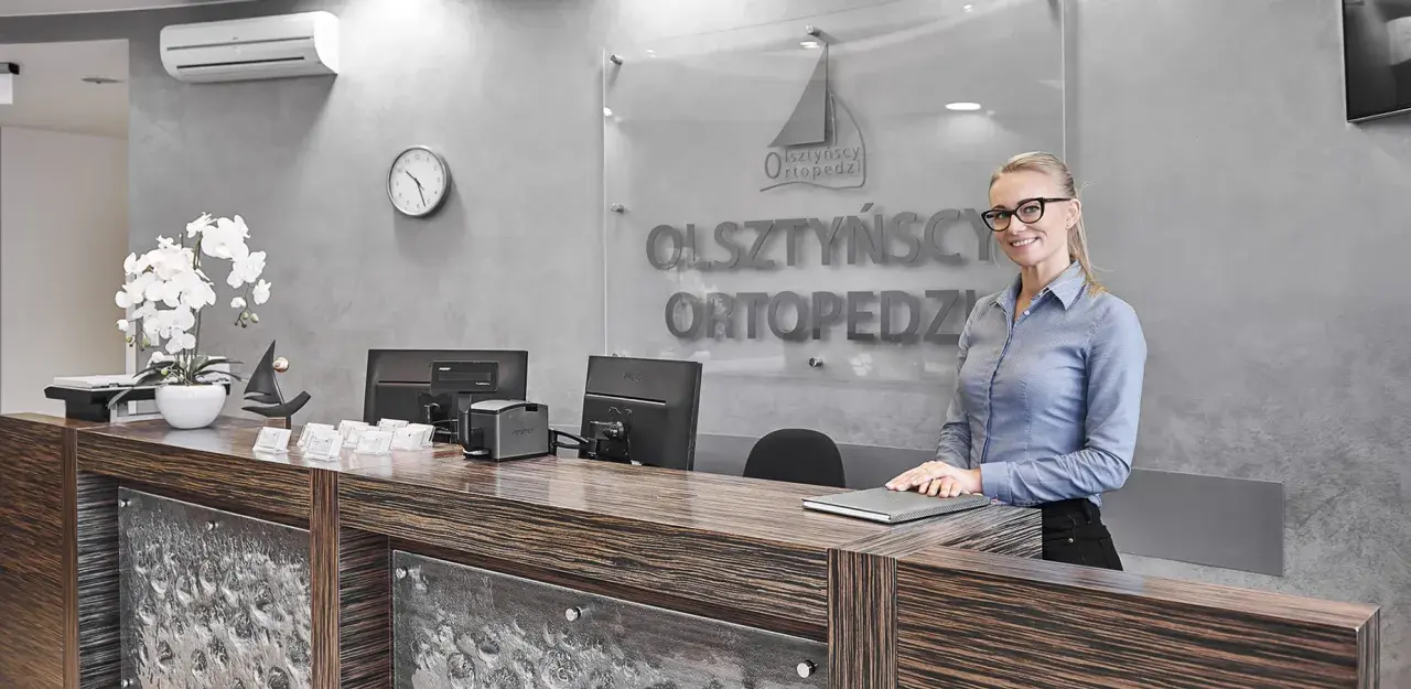 Recepcjonistka w okularach stoi przy ladzie w nowoczesnej klinice ortopedycznej.