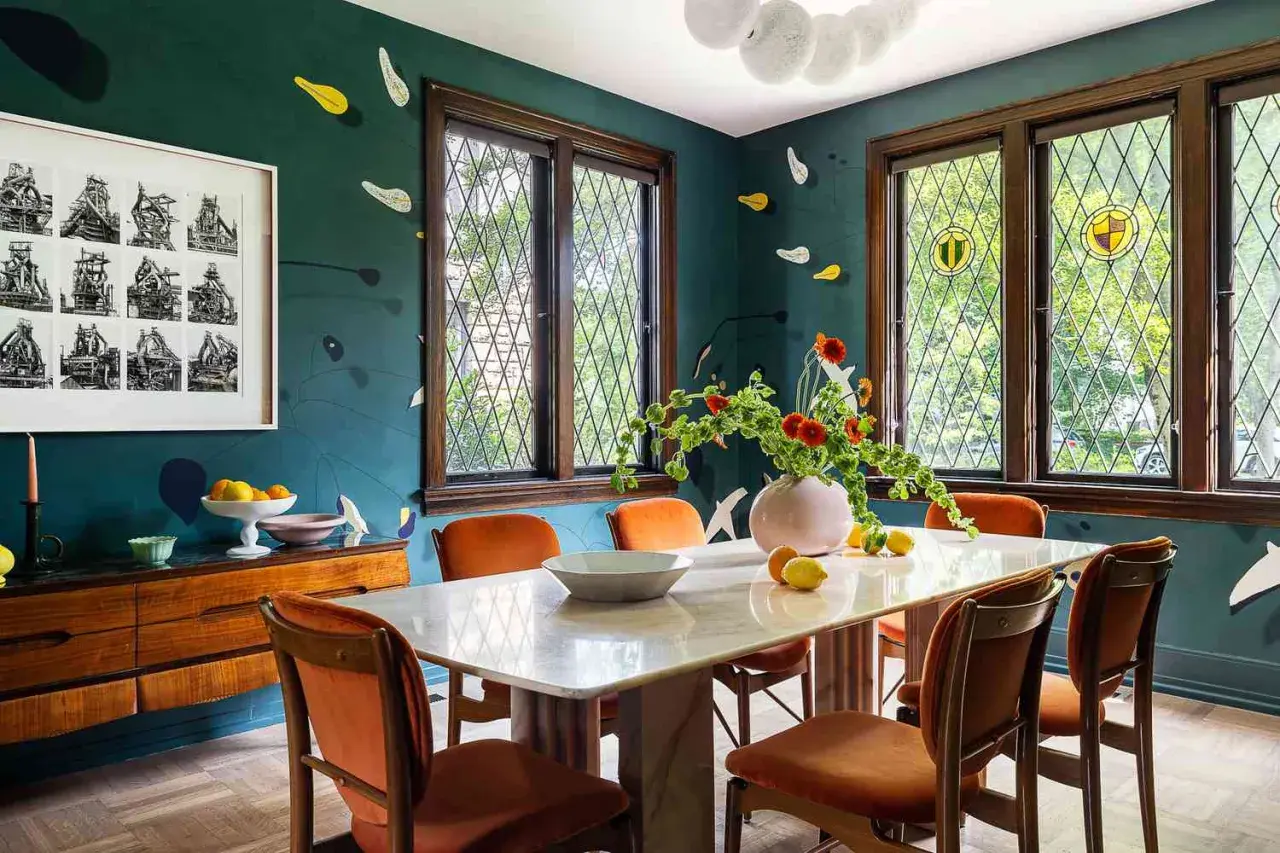 A vibrant kitchen and dining room design featuring a marble table, orange chairs, and a dark teal wall with playful accents.