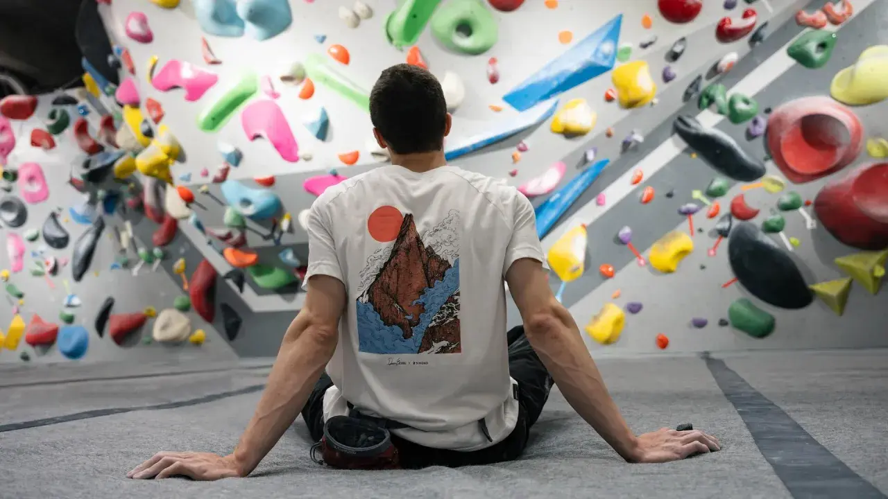 Mann sitzt vor einer Boulderwand mit bunten Griffen. Er ist bereit, besser klettern zu lernen.