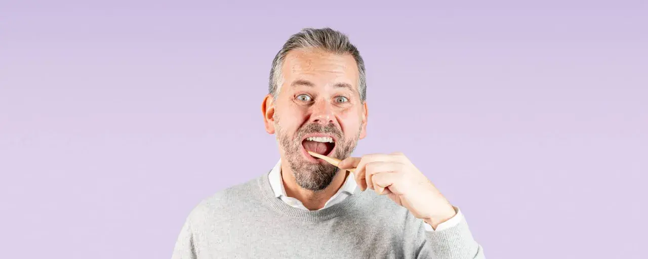 Uomo con barba che si lava i denti con uno spazzolino di legno, un gesto quotidiano per una corretta igiene dentale.