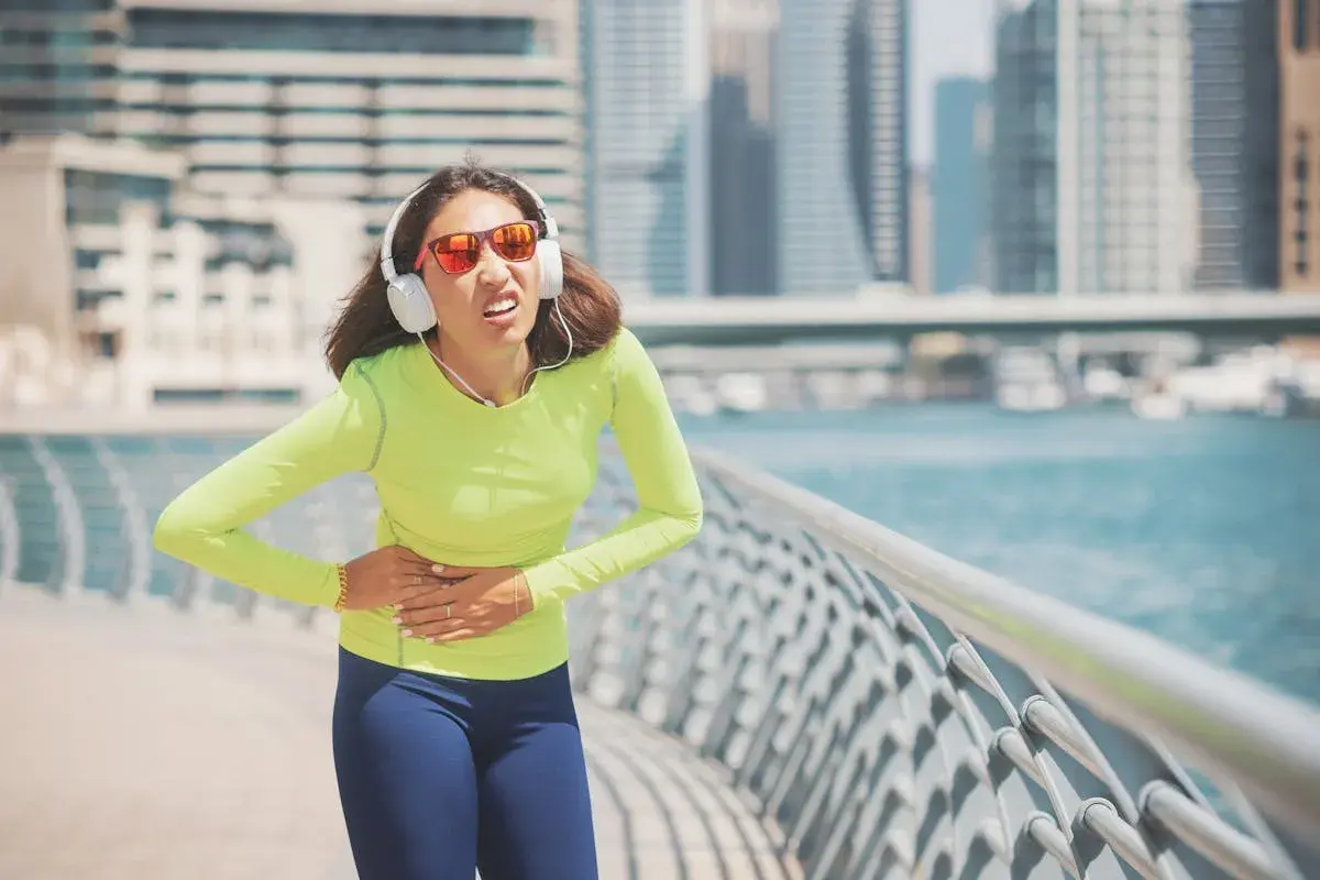 Une coureuse en tenue de sport, écouteurs aux oreilles, souffre d'un point de côté aigu en pleine course le long d'une promenade urbaine.