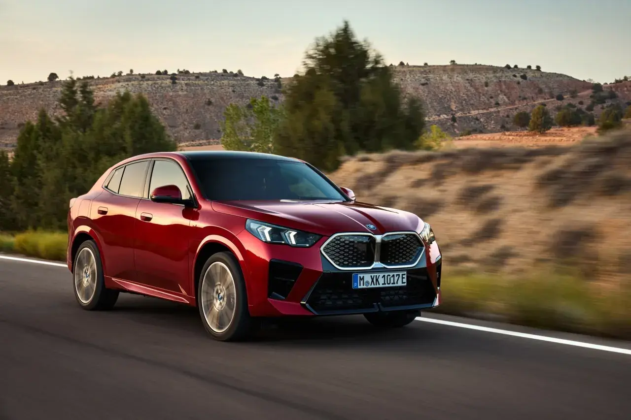 Czerwone BMW X2 2025 pędzi po pustynnej drodze, otoczone suchymi wzgórzami i drzewami.