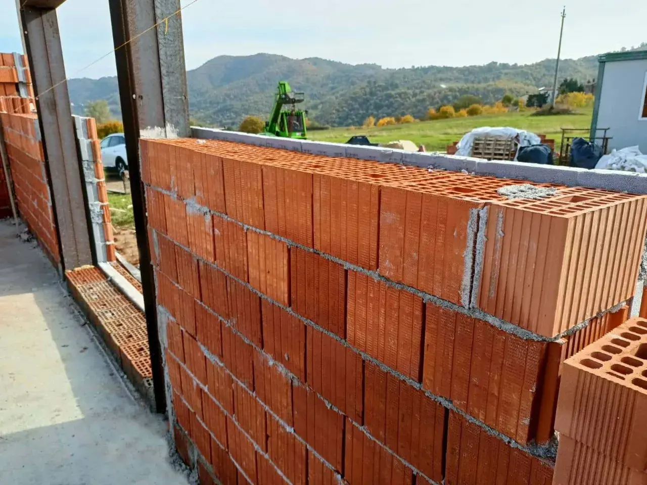Muro in costruzione con blocchi di laterizio, un materiale che resiste al fuoco, con vista su colline verdi e un escavatore.