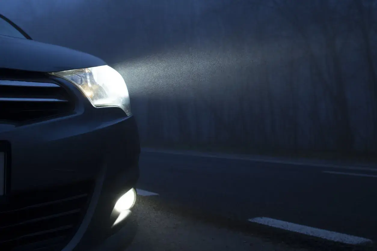 El coche avanza en la niebla con sus luces antiniebla encendidas, iluminando el camino oscuro y los árboles a un lado.