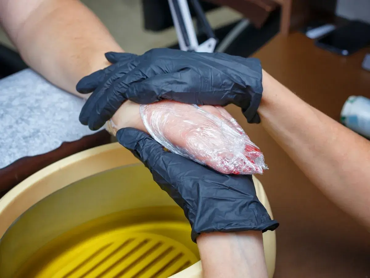 Dłonie w czarnych rękawiczkach owijają dłoń w folię, obok żółta wanna z płynem.