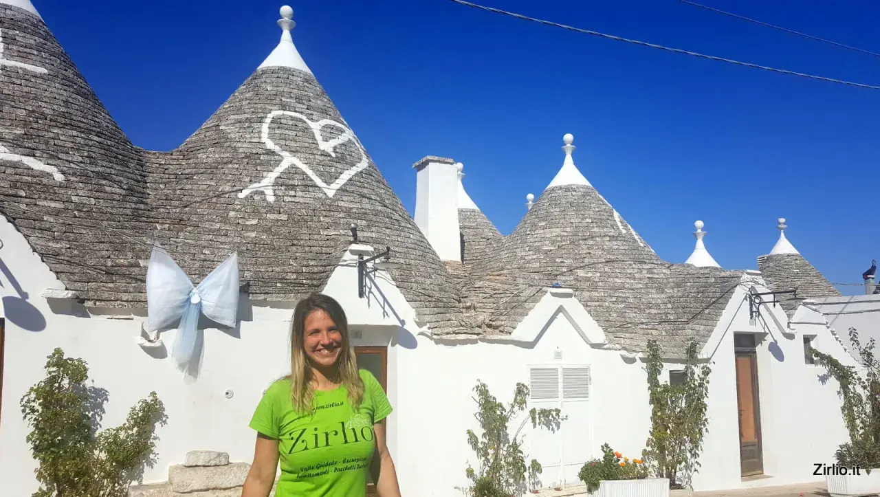 Una donna sorride davanti ai trulli di Alberobello, patrimonio UNESCO, con un grande fiocco azzurro appeso a un cono.