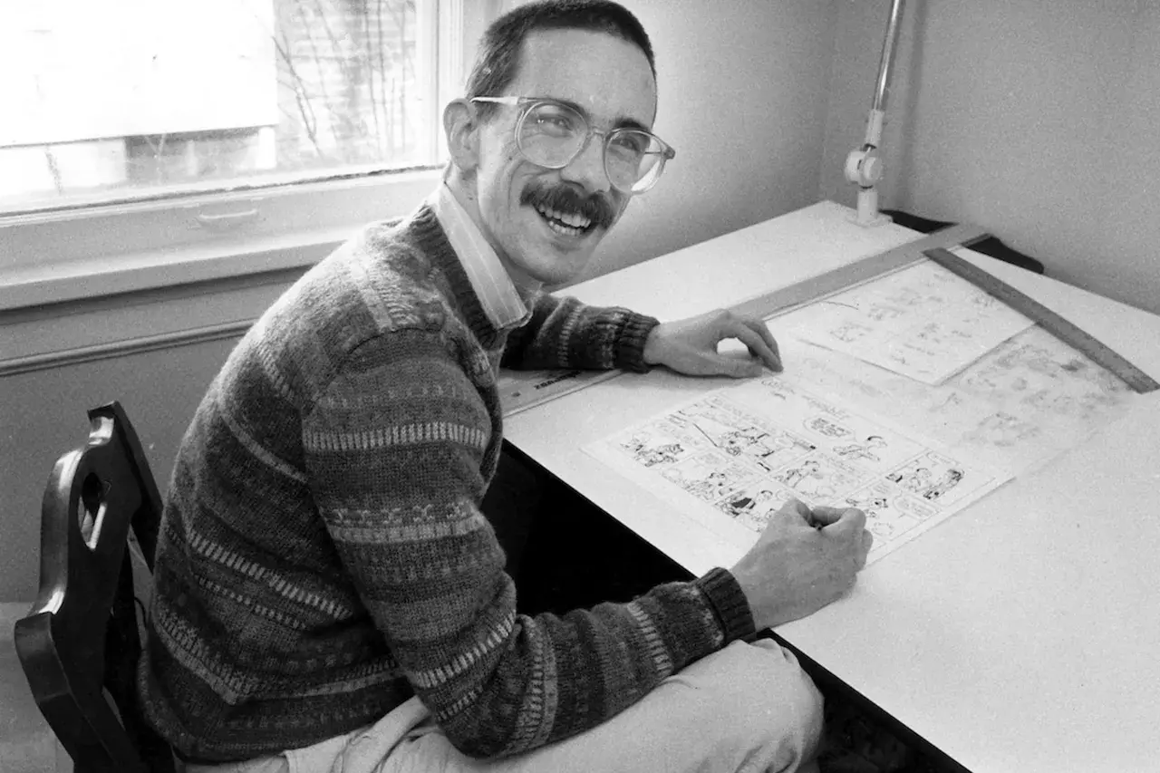 A smiling man with glasses and a mustache, likely Thomas Hobbes, draws a comic strip at his drafting table.