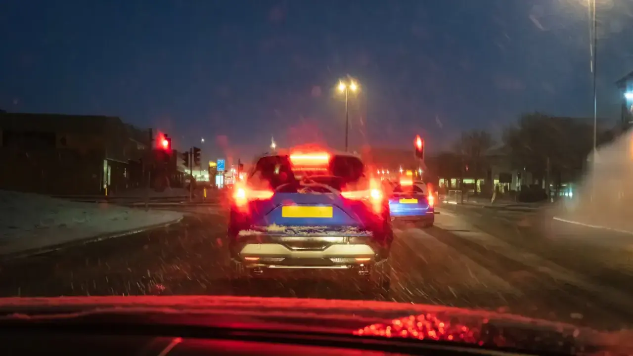 Noche de nieve, luces rojas de freno y un coche azul con matrícula amarilla. El asfalto mojado y la baja visibilidad aumentan el riesgo de derrape coche.