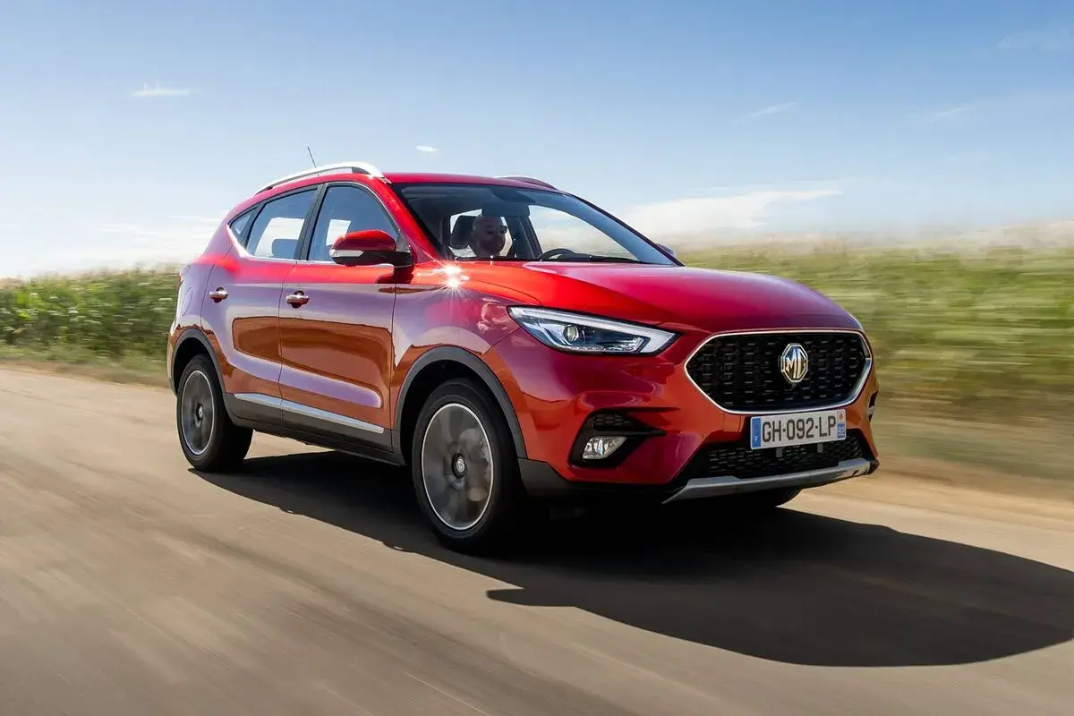 MG ZS en prueba, rojo brillante, circulando por un camino rural con campos verdes y cielo azul.