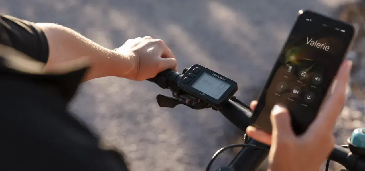 Rowerzysta trzyma kierownicę, na której zamontowany jest licznik rowerowy nie pokazuje prędkości. Obok wyświetlacz telefonu z połączeniem do Valerie.