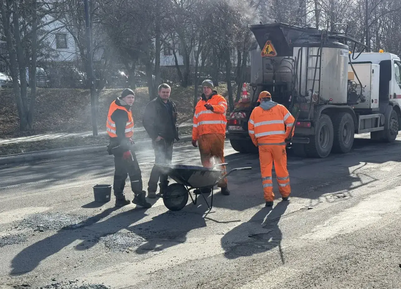 Ekipa drogowców w pomarańczowych strojach pracuje przy drodze, obok wózka i specjalistycznej ciężarówki.