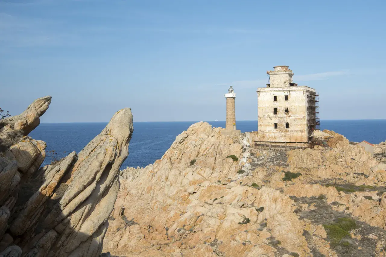 Il faro della Maddalena, con la sua torre e l'edificio adiacente in rovina, si erge su una costa rocciosa battuta dal mare.
