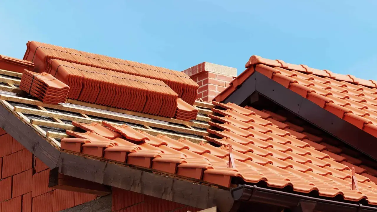 Układanie ceramicznych dachówek na dachu w budowie, widoczny komin i niebo.