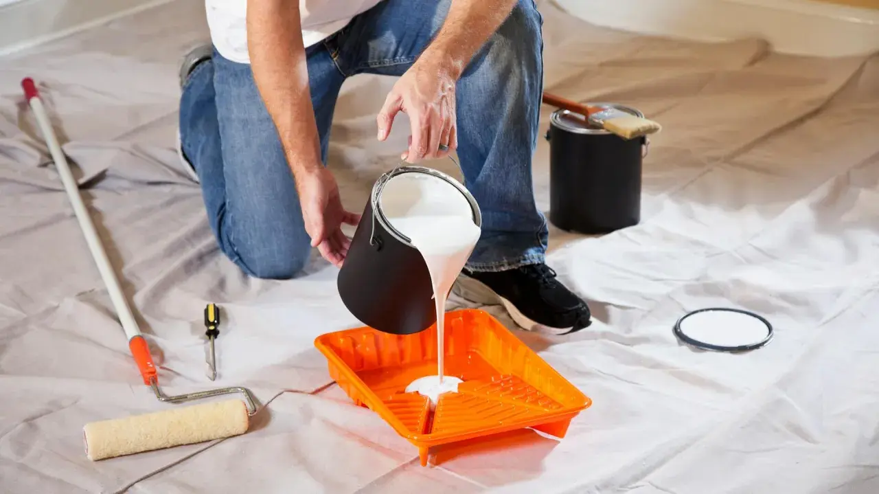 Uomo versa vernice bianca in una vaschetta arancione, pronto per pitturare soffitto con pennello. Attrezzi da pittura a portata di mano.