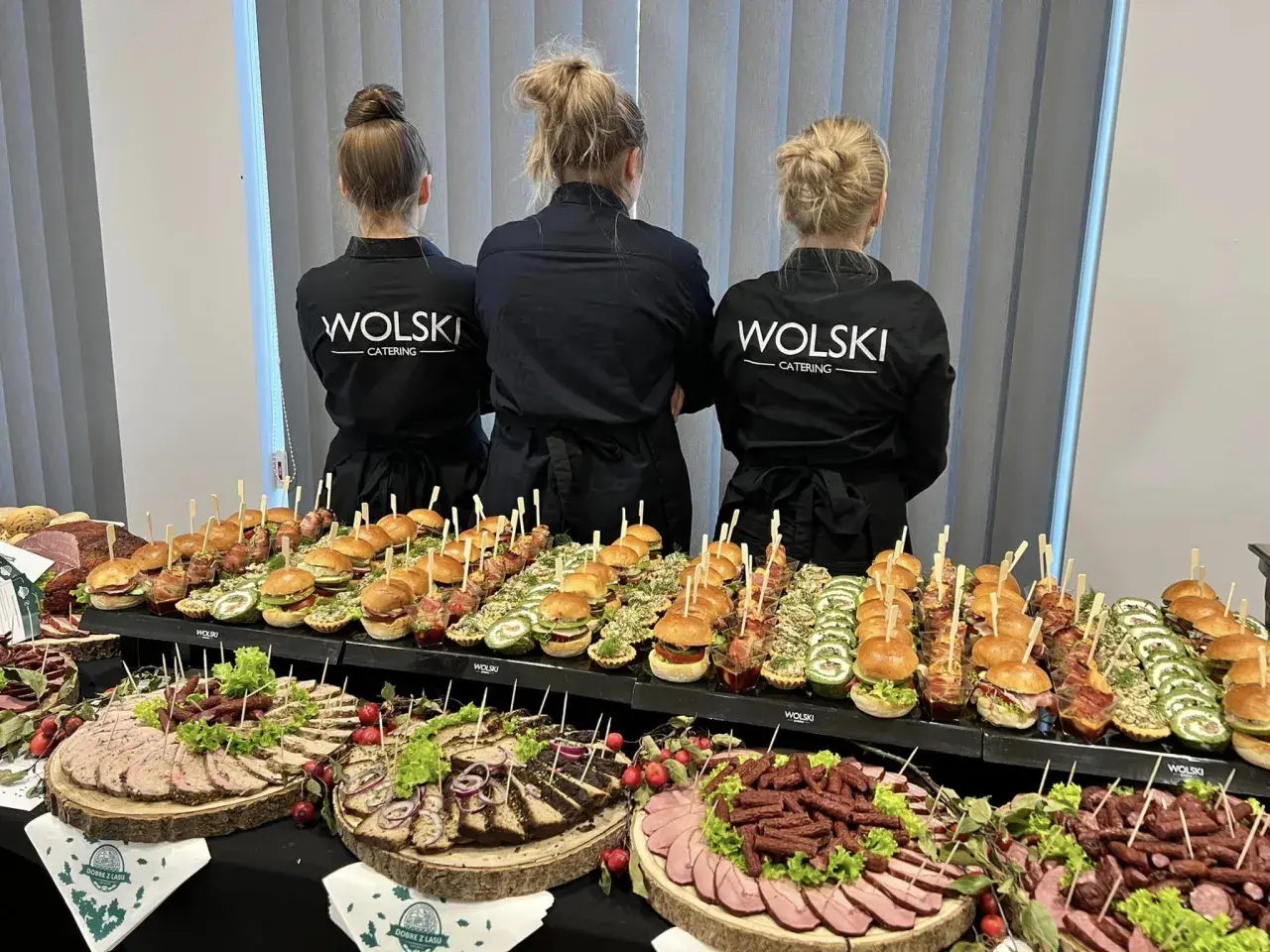 Trzy kobiety w czarnych fartuchach z logo "WOLSKI CATERING" stoją za stołem pełnym przekąsek. To idealna impreza z pysznym jedzeniem.