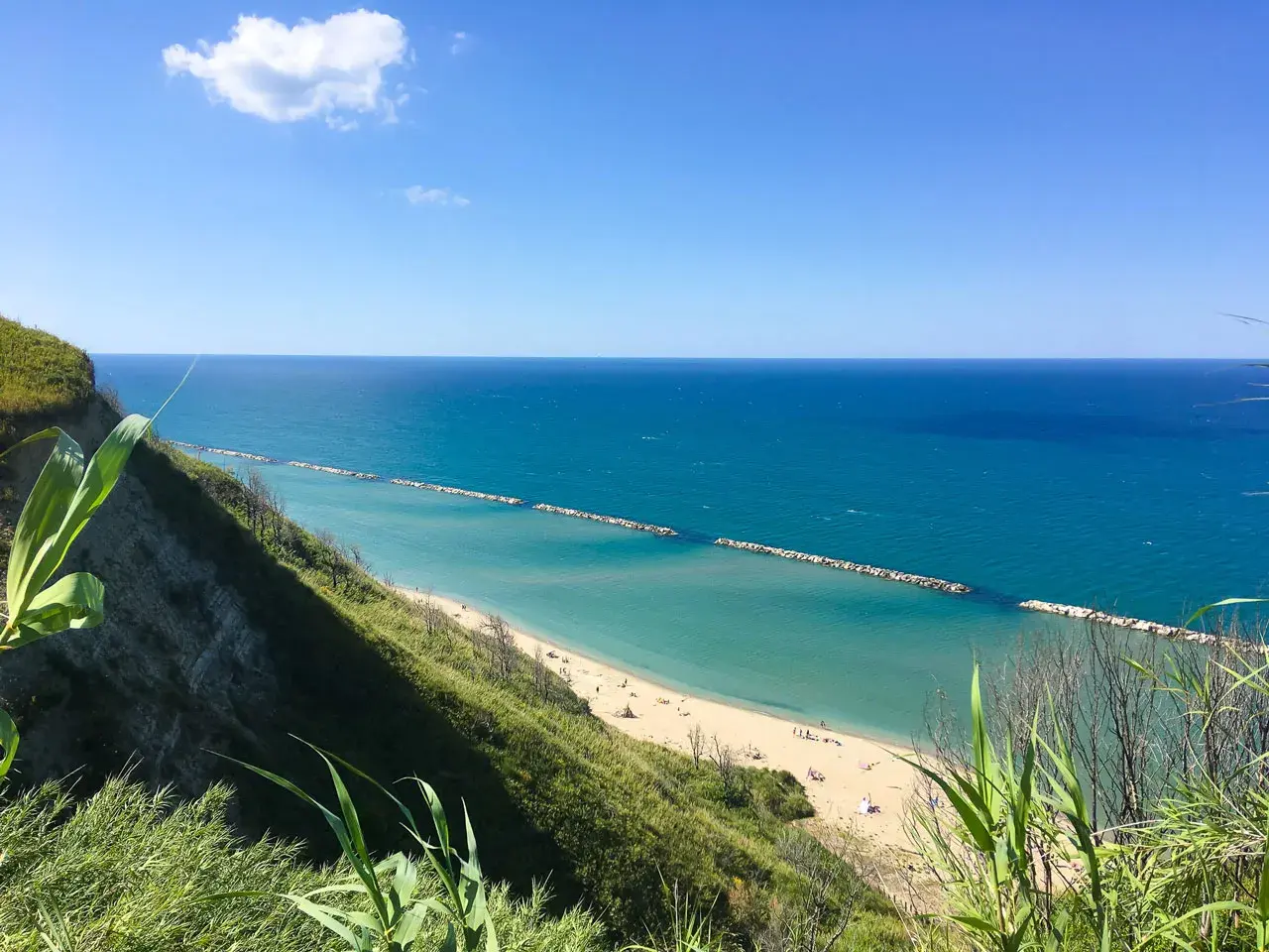 Vista panoramica della spiaggia di Pesaro con mare azzurro e scogliera verde. La focara è un ricordo lontano, ma la bellezza del paesaggio resta.