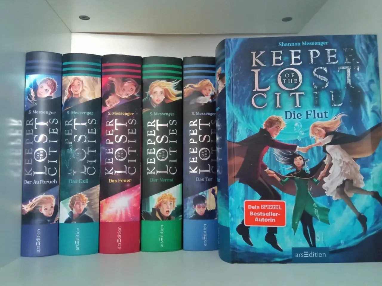 Die Bücherreihe "Keeper of the Lost Cities" mit Sophie als Hauptfigur. Mehrere Bände sind nebeneinander aufgestellt, der größte Band "Die Flut" steht rechts.
