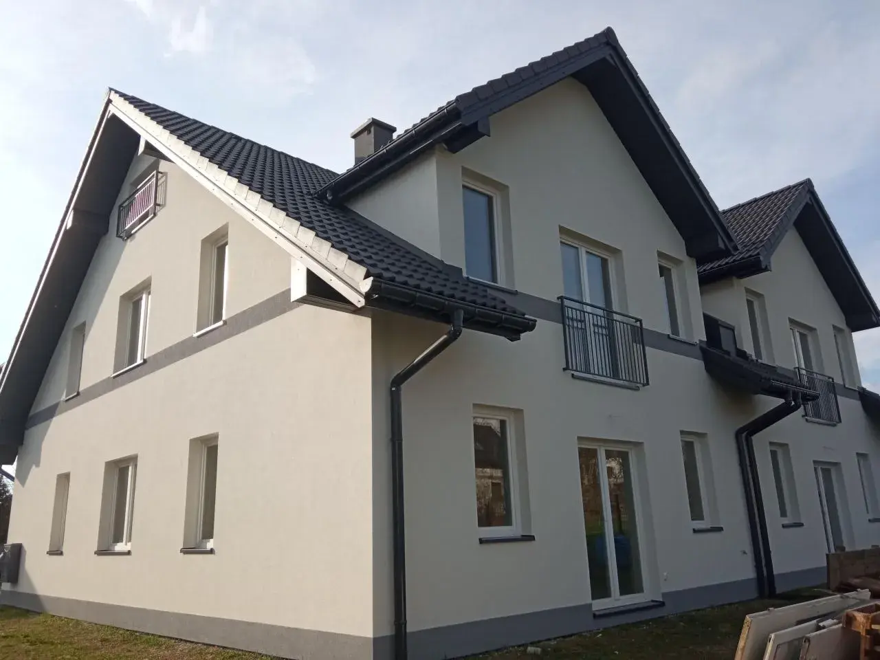 Nowy dom z jasną elewacją i ciemnym dachem. Wokół zielona rząska trawy, a na ścianach liczne okna i balkony.