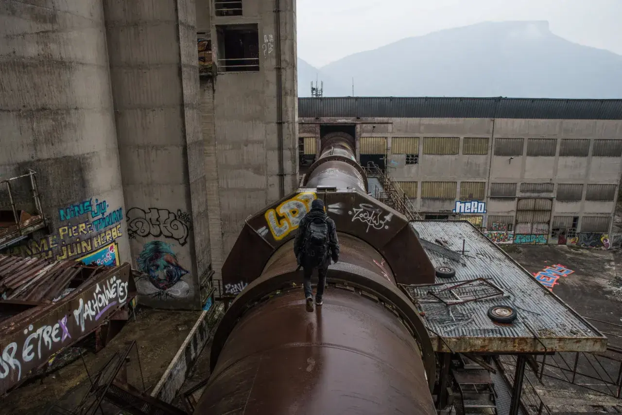 Explorateur en urbex, marchant sur un immense tuyau rouillé, traversant un grand vide industriel. Graffiti sur les murs.