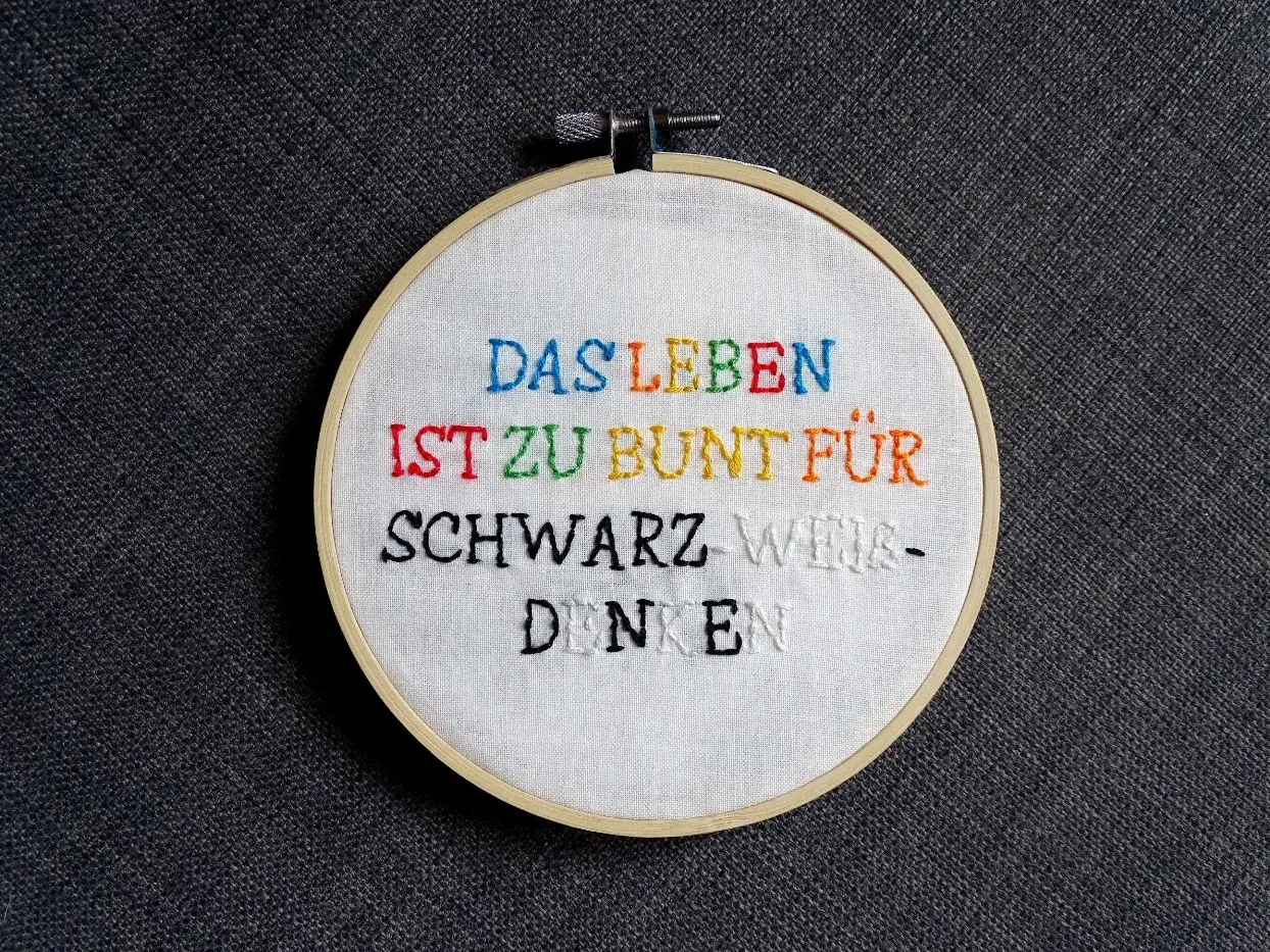 Ein Stickrahmen mit dem Spruch "Das Leben ist zu bunt für schwarz-weiß-denken", kunstvoll mit bunten **Buchstaben sticken** verziert.