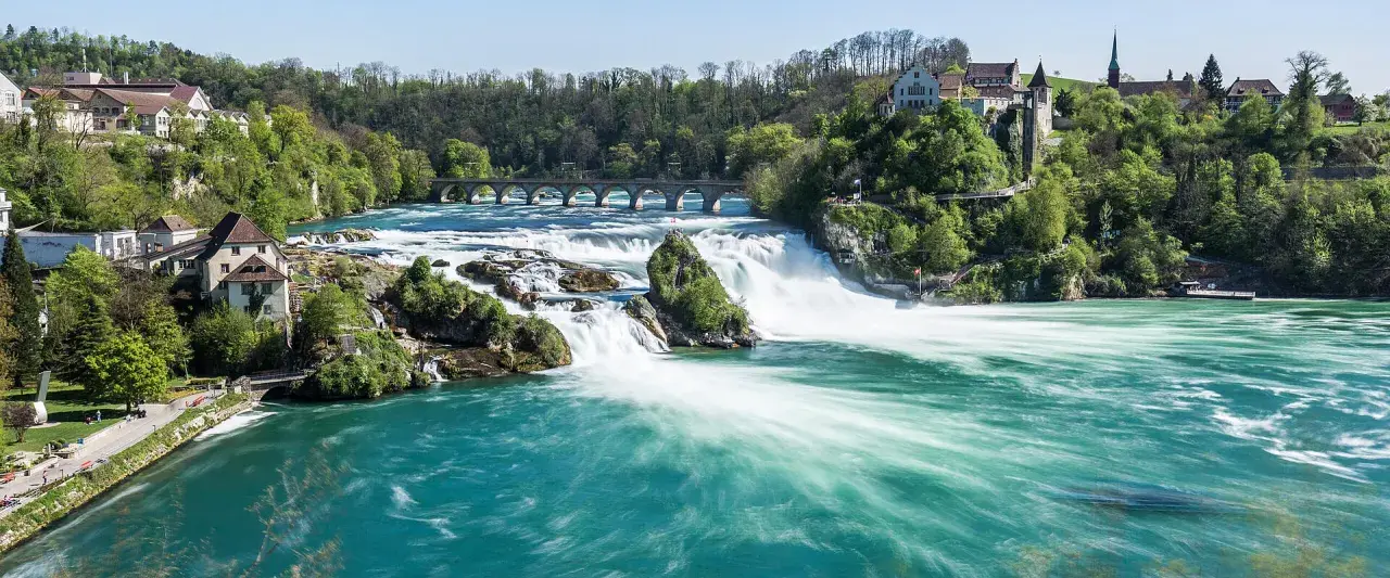 Breite, türkise Wasserströme stürzen über Felsen, umgeben von grünen Hügeln mit Häusern und einer Brücke. Die Rhine Waterfalls sind ein beeindruckendes Naturschauspiel.